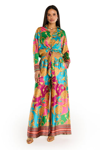 A woman stands confidently in sandals against a white background, wearing the Marcia Silk Pant - Dolce Sun: a vibrant designer set featuring a colorful baroque summer print with green, pink, blue, and gold on wide-leg silk pants and a long-sleeve knotted shirt.
