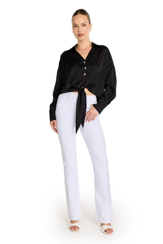 A woman stands against a white background wearing the Cecilia Silk Tie-Front Blouse in black, paired with white high-waisted flared pants, white heeled sandals, and hoop earrings.