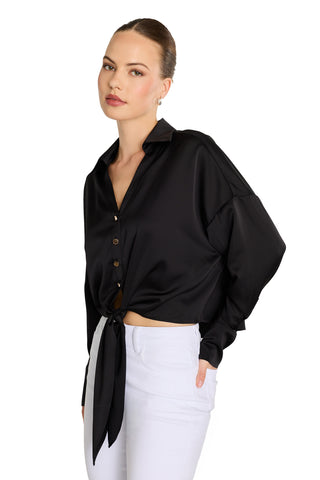 A woman poses against a white backdrop in the Cecilia Silk Tie-Front Blouse - Black, styled with high-waisted white pants and her hair pulled back, hand in pocket, highlighting the chic look of this stylish collared blouse.