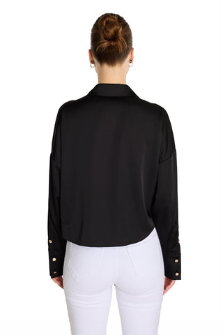 A woman with her hair in a neat bun is seen from the back, wearing the black Cecilia Silk Tie-Front Blouse and high-waisted white pants against a plain white background.