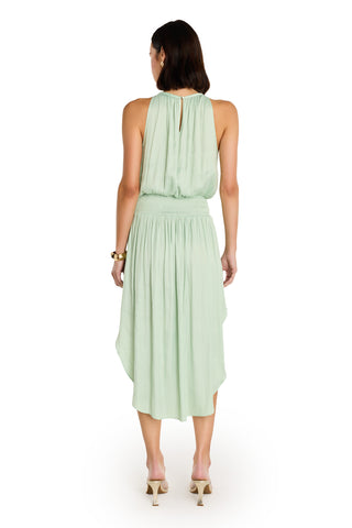A woman with shoulder-length dark hair models the Emelia Halter Dress in mint, featuring a sleeveless design, gathered waist, and uneven hem. She pairs it with heeled sandals and a bracelet against a white background.