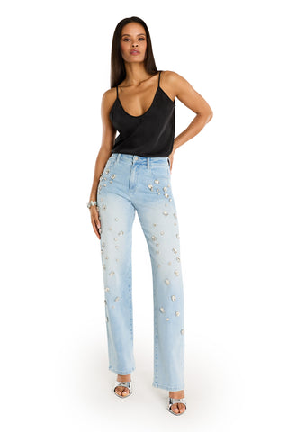 A woman poses confidently in a black satin camisole and Tara Crystal Jean - Light Blue high-rise jeans adorned with silver studs. She has straight brown hair, silver heels, and a bracelet, set against a plain white background.