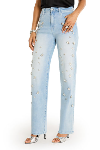 Someone wearing the Tara Crystal Jean - Light Blue, high-rise straight-leg jeans with large rhinestones, paired with a black top, silver high-heeled sandals, a bracelet, and rings, stands against a white background.