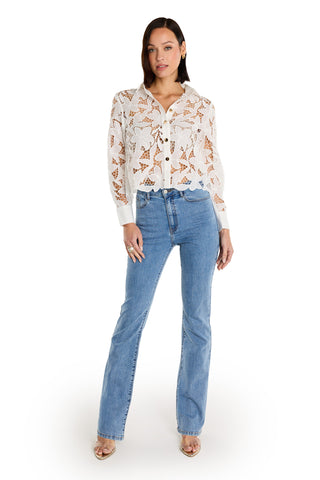 A woman with straight dark hair stands confidently against a white background, wearing the Mina Lace Blouse - White, blue high-waisted jeans, and open-toe heels.