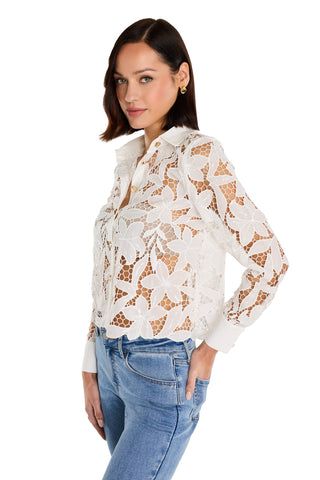 A woman with chin-length dark hair wears the Mina Lace Blouse - White, a sheer floral lace top, paired with light blue jeans. She stands sideways and smiles at the camera against a white background.