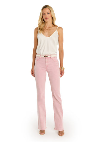 A woman with long blonde hair stands facing forward, wearing the Aria Flare Jean - Blush with a white sleeveless top and belt. She accessorizes with bracelets, necklaces, and nude heels against a white background.