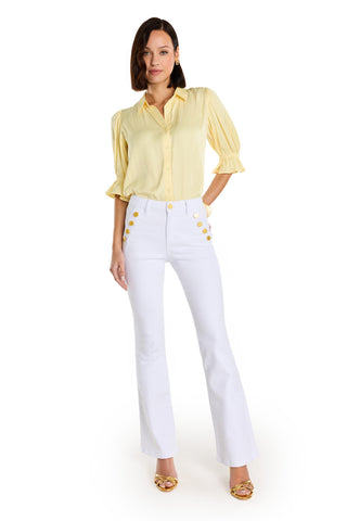 A woman stands facing forward in the Cindy Ruffle Sleeve Shirt - Sunshine Yellow and high-waisted white flared pants with gold buttons, paired with gold open-toe heels. She is set against a white background.