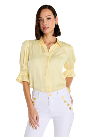 A woman with medium-length dark hair wears the Cindy Ruffle Sleeve Shirt in Sunshine Yellow and high-waisted white pants with large gold buttons, standing against a plain white background.