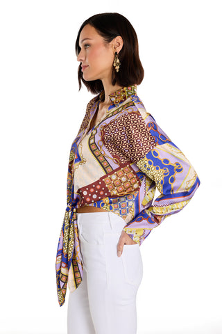 A woman with chin-length dark hair and dangling gold earrings stands in profile, hand in pocket, wearing the Cecilia Silk Tie-Front Blouse in Riviera Chain with gold accents and white pants, posing against a white background.