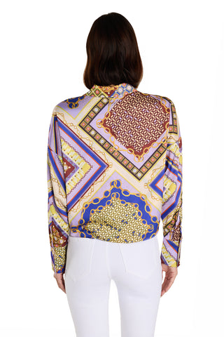 A woman with medium-length brown hair, seen from behind, wears the Cecilia Silk Tie-Front Blouse in Riviera Chain, featuring geometric and ornate patterns in purple, gold, blue, and red, paired with high-waisted white pants.