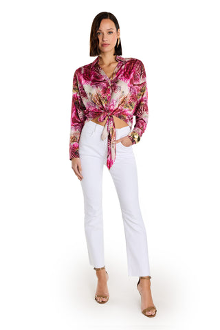 A woman stands against a white background wearing the Cecilia Silk Tie-Front Blouse in Bright Orchid with a pink and white floral print, paired with white high-waisted pants, gold heels, and a gold watch. Her hair is styled in a side part.