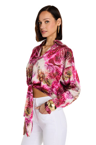 A woman with straight brown hair wears the Cecilia Silk Tie-Front Blouse - Bright Orchid, a pink and white floral silk top, styled with white pants and gold accessories, posing against a white background.