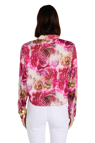 A person with shoulder-length brown hair is shown from the back, wearing the Cecilia Silk Tie-Front Blouse in Bright Orchid, featuring a silky light-weight fabric with a pink, white, and yellow abstract pattern, paired with high-waisted white pants.