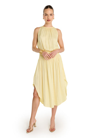 A woman stands against a white background wearing the Emelia Halter Dress in sunshine yellow, featuring a sleeveless design, gathered waist, and asymmetrical hem. She pairs it with strappy heeled sandals and has her hair pulled back, hands gently clasped.