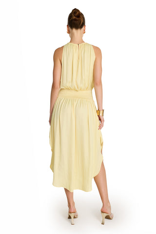 A woman faces away, wearing the Emelia Halter Dress in sunshine yellow, a sleeveless midi with gathered waist and asymmetrical hem, styled with heeled sandals and a gold cuff bracelet against a white background.