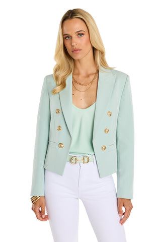 A woman with long blonde hair wears a mint Gabrielle Double Breasted Blazer with gold buttons over a matching top, white pants, and gold jewelry, standing against a plain white background.