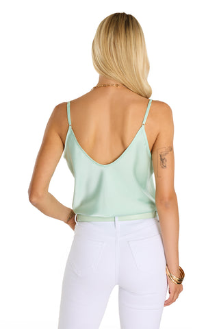 A woman with long blonde hair, wearing the Yara Silk Camisole in mint with adjustable straps and white pants, stands with her back to the camera. She has a tattoo on her upper left arm and wears gold bracelets and a necklace.