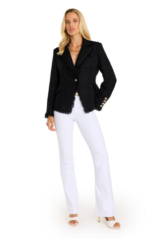 A woman stands confidently in the Giselle Single Breasted Tweed Blazer - Black with gold buttons, paired with white flared pants, white open-toe heels, layered necklaces, and long blonde hair.