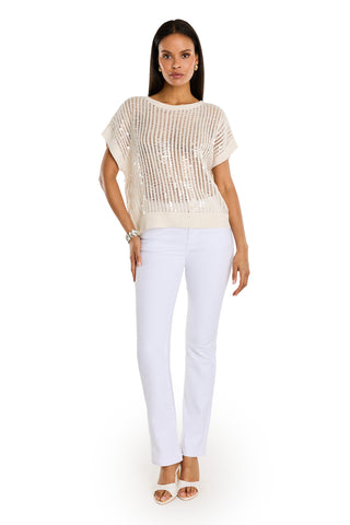 A woman with long dark hair wears the Carmela Sequin Knit Top in pearl over a white tank, with white pants, open-toe heels, and a bracelet while facing forward.