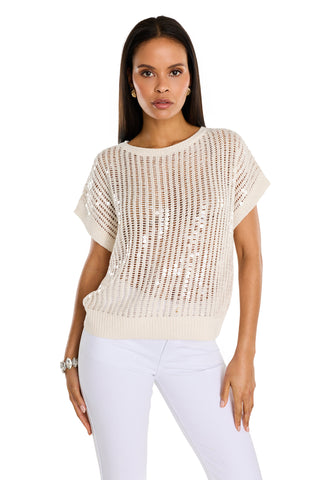 Woman wearing the Carmela Sequin Knit Top in pearl over a white top and white pants, standing with one hand on her hip against a plain white background.