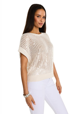 Woman with long dark hair wearing the Carmela Sequin Knit Top in Pearl over a light top, paired with white pants and a bracelet, standing against a white background.