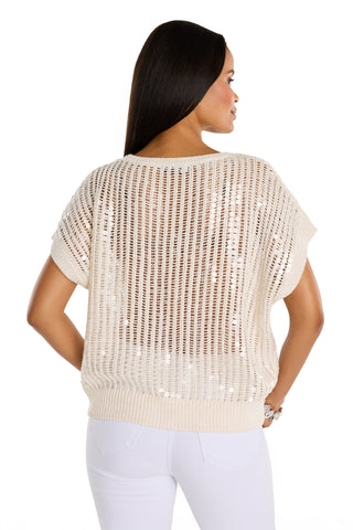 A woman with long dark hair faces away, wearing the Carmela Sequin Knit Top in pearl over a white top and white pants, set against a plain white background.