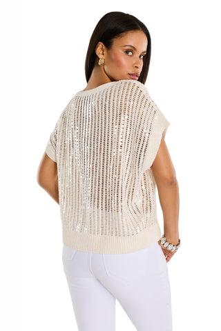 A woman with straight dark hair models the Carmela Sequin Knit Top in pearl, featuring an open-knit, short-sleeve design with sequins. She pairs it with white pants and poses facing backward over her shoulder against a white background.