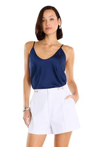 A woman stands against a white background with one hand in her pocket, wearing a dark blue sleeveless satin top and the Ellery Horsebit Short in white, featuring gold horsebit hardware.