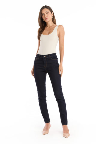 A woman stands against a white background, wearing a sleeveless beige bodysuit, nude heels, and the Layla Skinny Jean - Indigo. She poses with one leg slightly bent and her arms relaxed by her sides.