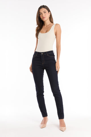 A woman with long brown hair wears the Loren Bodysuit in Pearl, dark blue skinny jeans, and nude heels, standing against a plain white background.