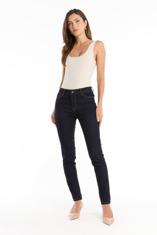 A woman stands against a white background, wearing a sleeveless beige top, Layla Skinny Jean - Indigo, and nude heels, with her hair down and one hand in her pocket.