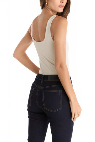 A woman faces away, wearing the Loren Bodysuit in Pearl paired with dark blue jeans featuring yellow stitching. Her right arm is bent, and her head is slightly turned to the side.
