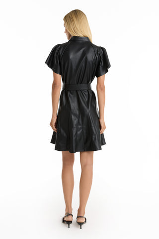 A woman with long blonde hair, seen from behind, wears the short-sleeve black Devin Leather Ruffle Dress with a waist belt and pleated skirt, paired with black heeled sandals, against a plain white background.