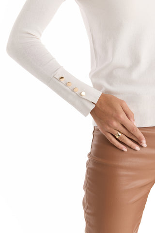 A person wears the Marcela Knit Top - Pearl, a fitted cream sweater with gold cuff buttons, paired with tan leather pants and a gold ring on one hand resting at their hip. The background is white.