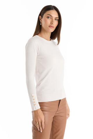 A woman with straight brown hair models the Marcela Knit Top - Pearl, a white fitted long-sleeve sweater with gold cuff buttons, paired with tan leather pants against a plain white background.