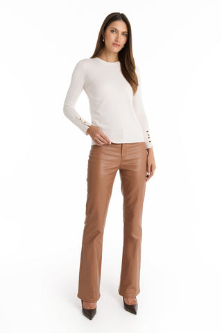A woman with long brown hair wears the Marcela Knit Top - Pearl, a fitted sweater with gold buttons, paired with high-waisted brown leather pants and brown pointed heels, standing against a white background.