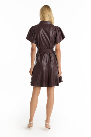 A woman with long, straight blonde hair stands facing away, wearing the Devin Leather Ruffle Dress in Merlot and white heeled shoes against a plain white background.