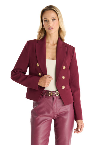 A woman wears the Gabrielle Double Breasted Blazer in Merlot with gold buttons and matching burgundy leather pants, posing with one hand on her blazer over a cream-colored top.