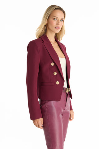 A woman stands against a white background, wearing the Gabrielle Double Breasted Blazer - Merlot with gold buttons over a white top and matching burgundy pants, looking slightly to the side.