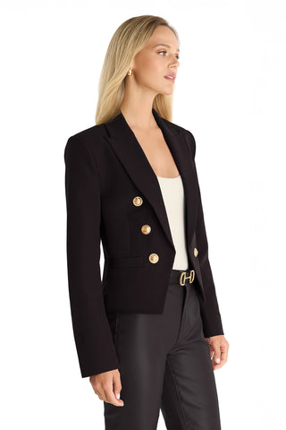 A woman with long blonde hair wears the Gabrielle Double Breasted Blazer - Black with gold buttons over a white top and black pants, standing in a three-quarter pose against a white background.