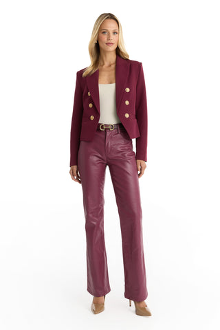 A woman with blonde hair and a confident expression wears a Gabrielle Double Breasted Blazer - Merlot with gold buttons, matching leather pants, a white top, and tan heels against a white background.