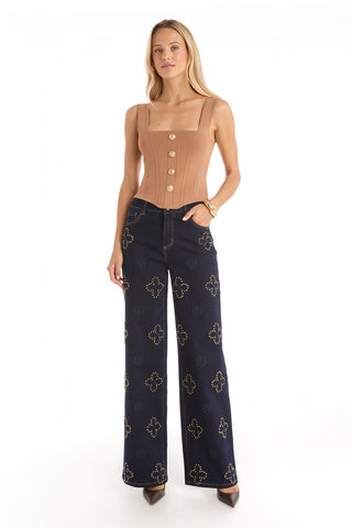 A woman with long blonde hair and brown heels stands against a white background wearing the Venus Button Top - Mocha Mousse, a tan, corset-inspired, sleeveless top with large buttons, paired with high-waisted dark blue embellished wide-leg jeans.