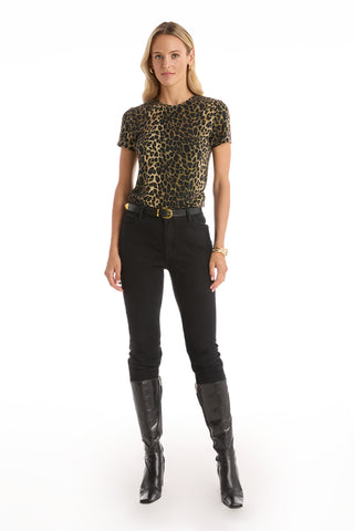 A woman with blonde hair faces forward against a plain white background, wearing the Amaya Ultra-Soft T-Shirt in leopard print, black belted pants, and knee-high black boots, with minimal accessories.