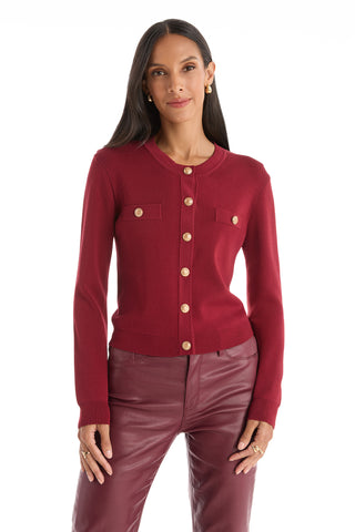 A woman stands facing forward against a white background wearing the Loretta Knit Cardigan Top in merlot with padded shoulders, chest pockets, gold buttons, and long sleeves, paired with high-waisted burgundy leather pants.