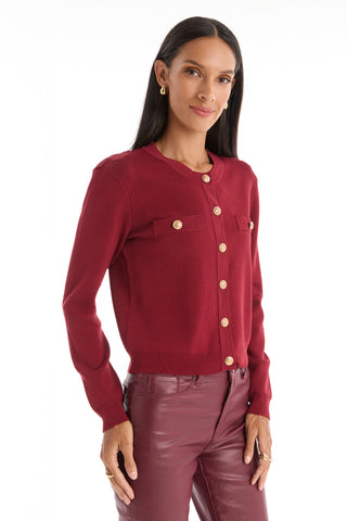 A woman with long dark hair wears the Loretta Knit Cardigan Top in Merlot, featuring padded shoulders, front pockets, and gold buttons. She pairs it with maroon leather pants and smiles softly against a white background.