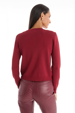 A woman with long dark hair wears the Loretta Knit Cardigan Top in Merlot, featuring gold buttons, paired with matching red leather pants, and stands facing away from the camera against a white background.