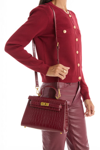 A woman in a red button-up sweater and red leather pants holds the Kayla Croco Handbag - Merlot, featuring a dark red crocodile texture, gold clasp, and shoulder strap.