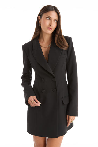 A woman with long brown hair wears the Kira Blazer Dress in black, a fitted, double-breasted design with power shoulders and long sleeves, styled with gold jewelry as she poses against a white background.
