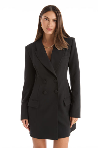 A woman with straight brown hair wears the Kira Blazer Dress in black, featuring a double-breasted design, power shoulders, and flap pockets, standing against a plain white background.