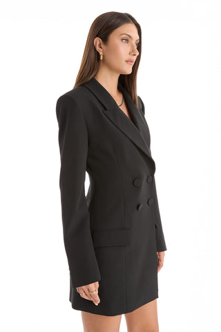 A woman with long brown hair stands against a white background, wearing the Kira Blazer Dress in black, featuring power shoulders, double-breasted buttons, long sleeves, and notch lapels. She faces slightly right with a neutral expression.